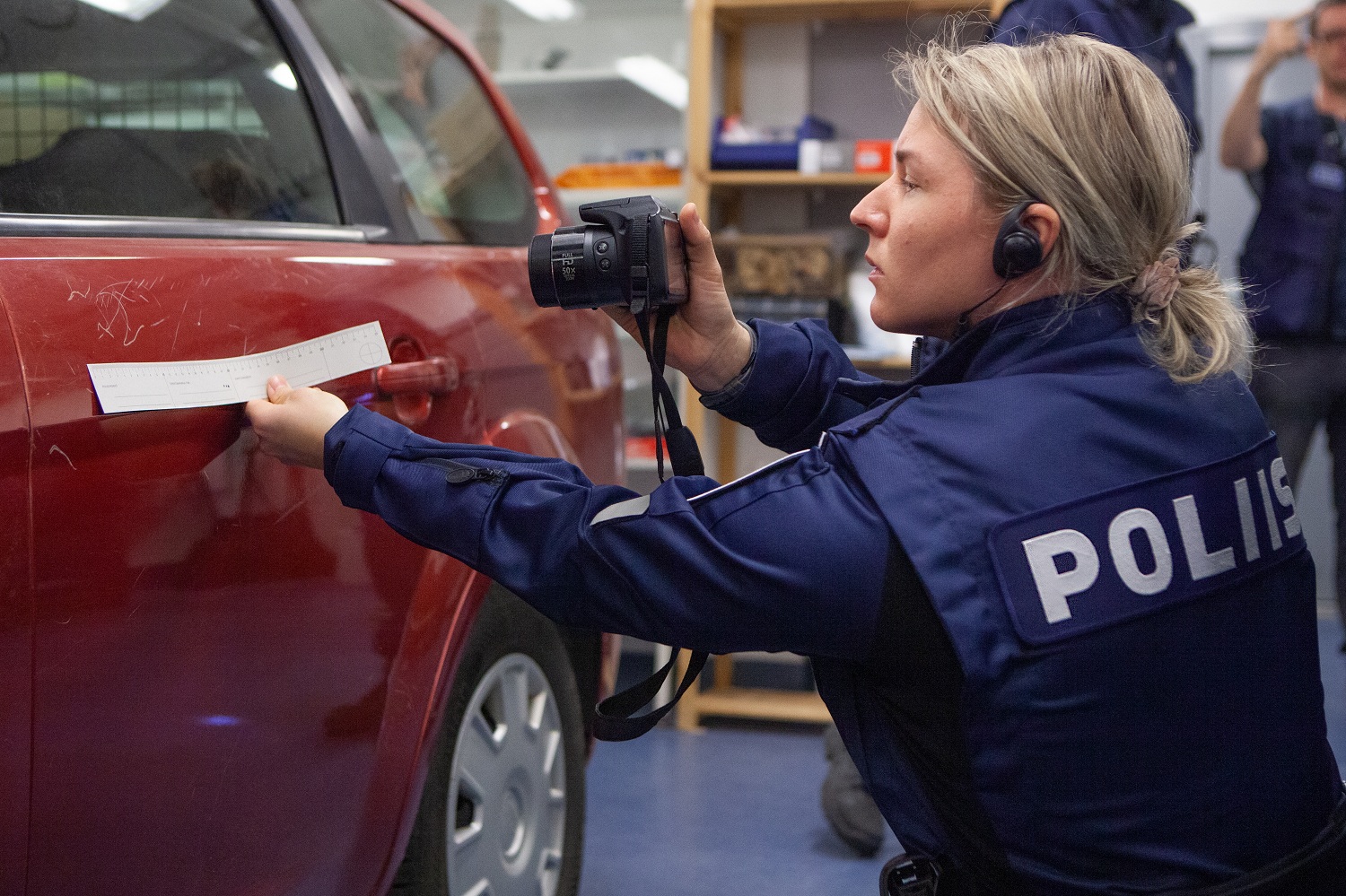 Virkapukuinen opiskelija ottaa kuvaa punaisen auton oven naarmuista.