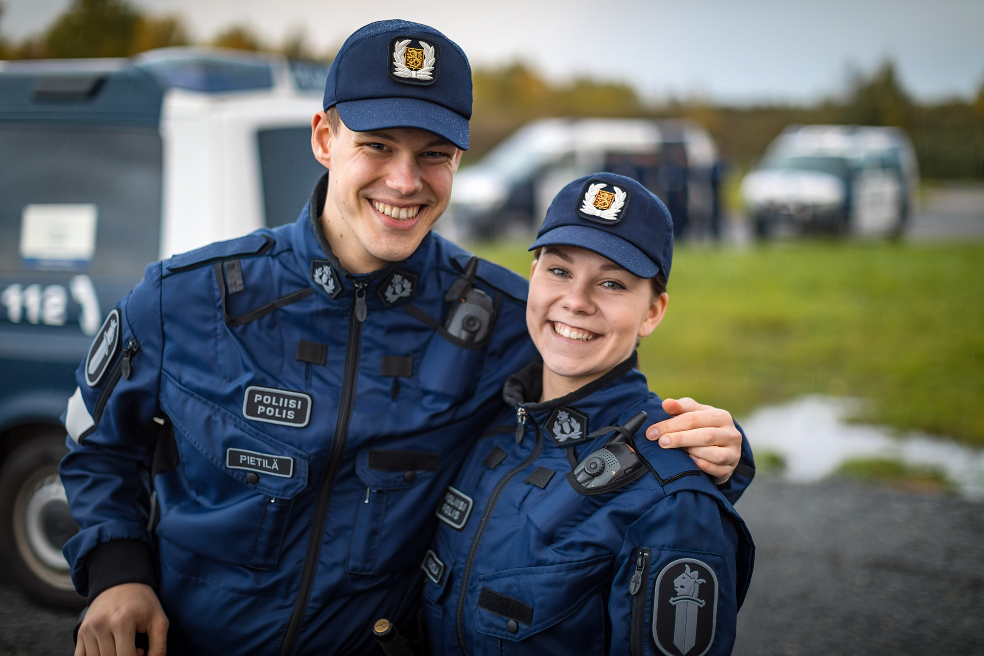 Kaksi virkapukuista poliisiopiskelijaa hymyilee, taustalla poliisiautoja.