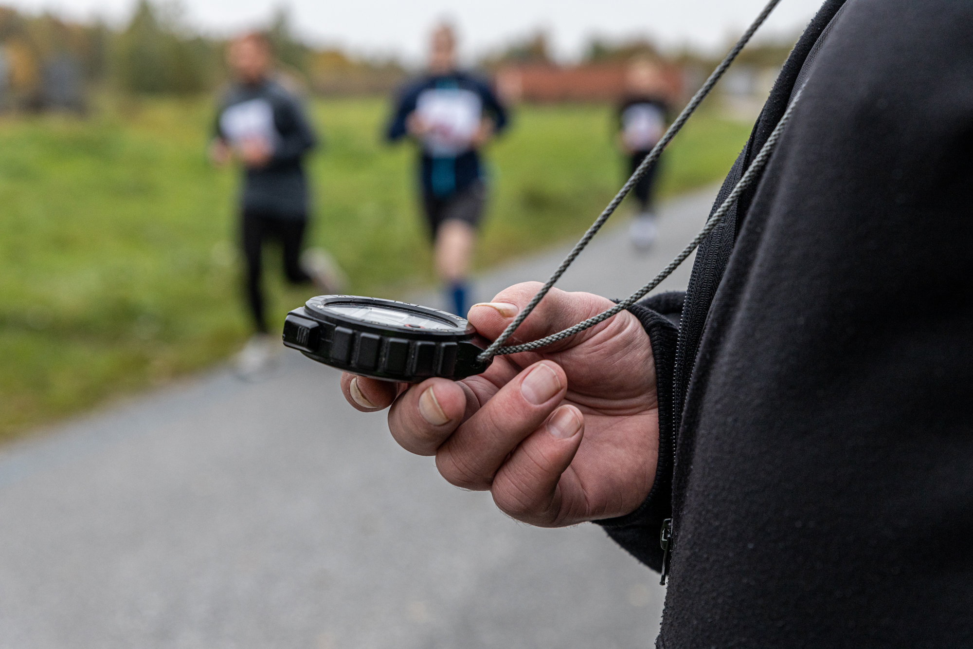 Ett tidtagarur i handen på en övervakare av löptestet, i bakgrunden syns sökande som springer.