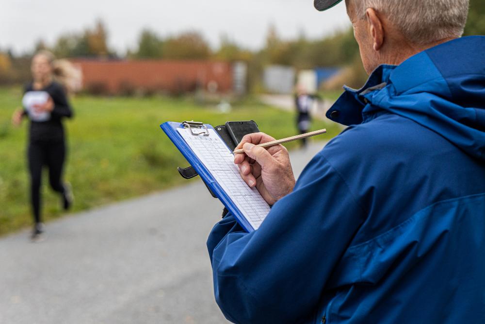 En övervakare av löptestet gör anteckningar på ett papper, i bakgrunden syns sökande som springer.