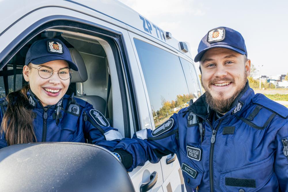 Kaksi hymyilevää poliisiopiskelijaa poliisiauton luona, toinen auton sisällä, toinen auton vieressä.