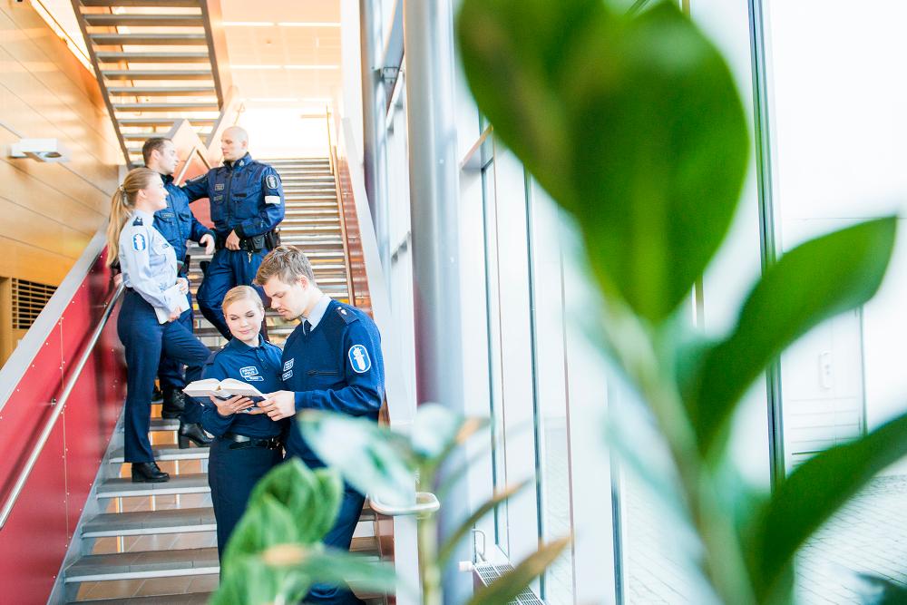 Två polisstuderande i uniform står i en trapp och läser en bok. I bakgrunden står tre studerande i uniform och pratar.