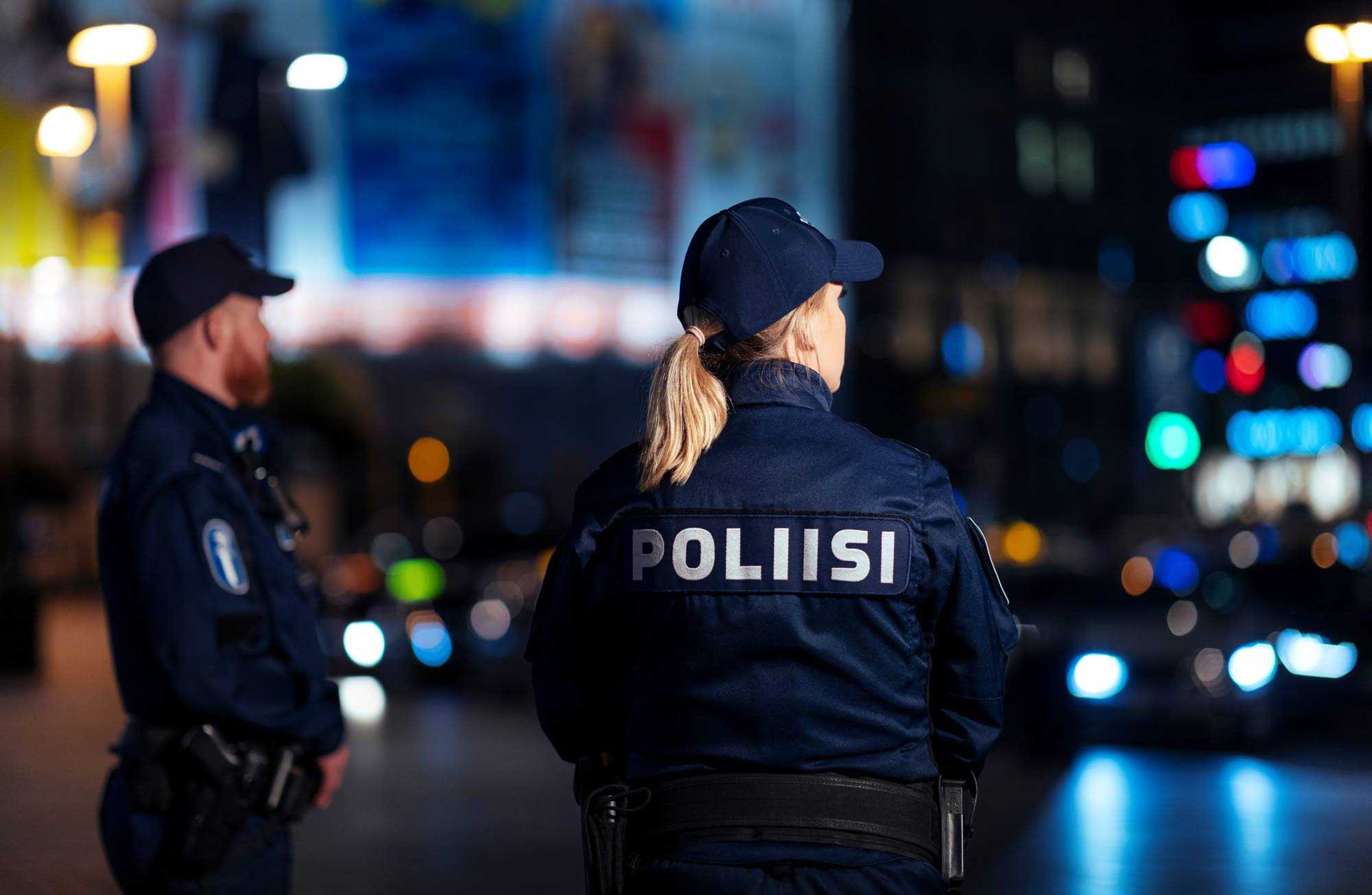 Två poliser i uniform ser sig omkring, runt dem en stad i kvällsljus. 