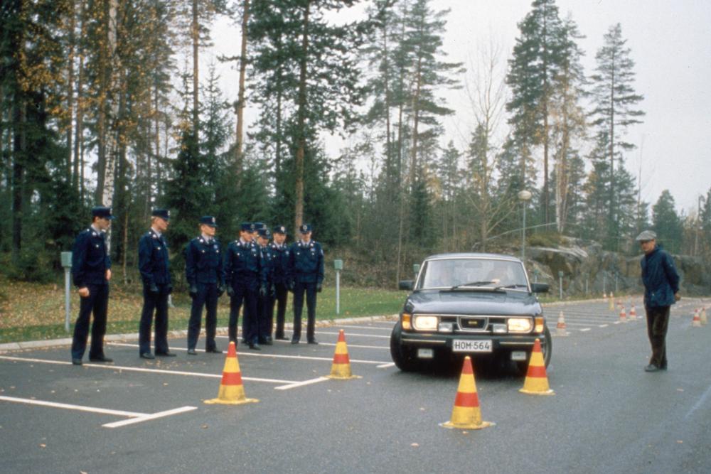 Övningskörning på Polisskolans parkeringsområde på 1980-talet. Åtta poliser i uniform och utbildaren tittar på när en Saab kör mellan konerna.