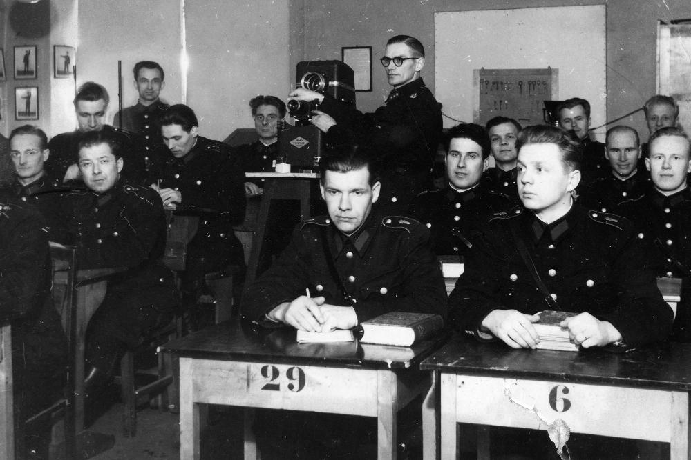 Uniformerade studerande på Rörliga polisens nybörjarkurs sitter i pulpeter på 1950-talet. Läraren justerar förstoringsanordningen för fotografier.