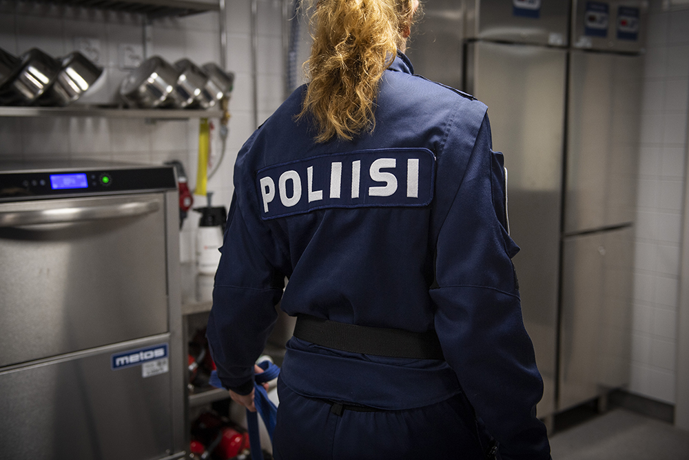 Ryggen av en polis i uniform som står i köket, på ryggen syns texten POLIISI. I köket finns bland annat matkoppar för hundar och köksmaskiner.