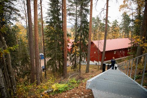  In forest scenery, there is a red building and stairs, where a uniformed police officer is walking with a dog.