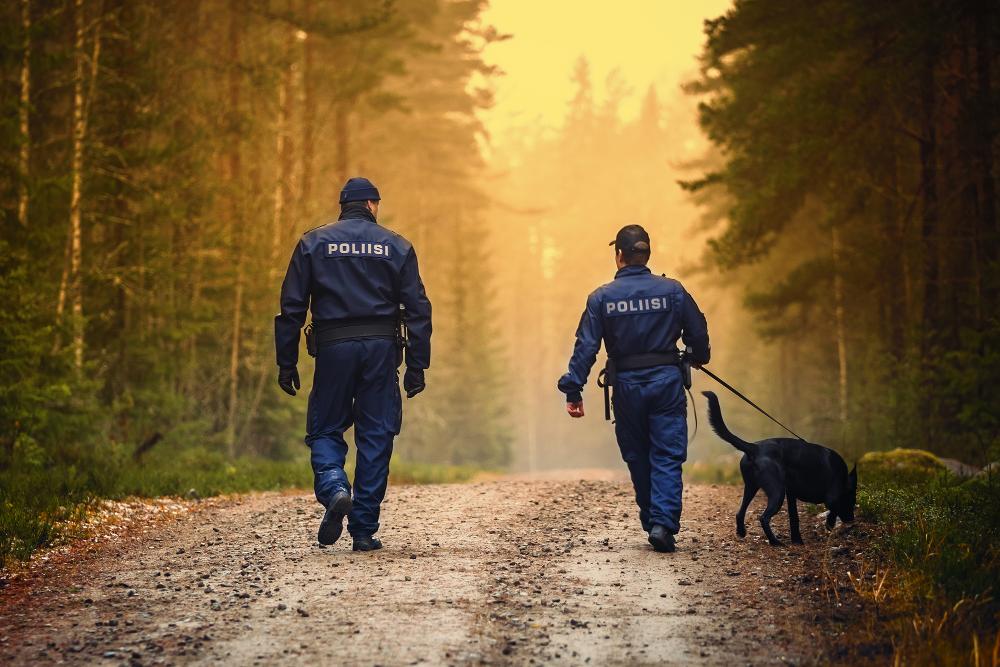 Kaksi virkapukuista poliisia kävelee talutushihnassa olevan koiran kanssa metsätiellä.