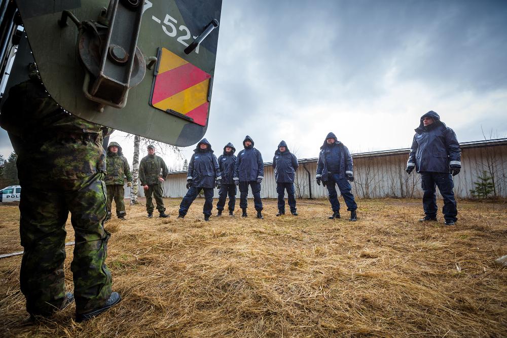 I förgrunden ligger en person i kamouflagekläder bakom dörren till ett bepansrat fordon i kamouflagefärger. Framför honom syns personer klädda i blå uniformer och kamouflagekläder.