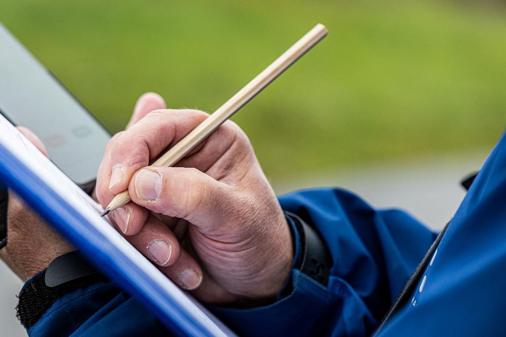 A hand holding a pencil is writing in a notepad. Green grass is visible in the background.