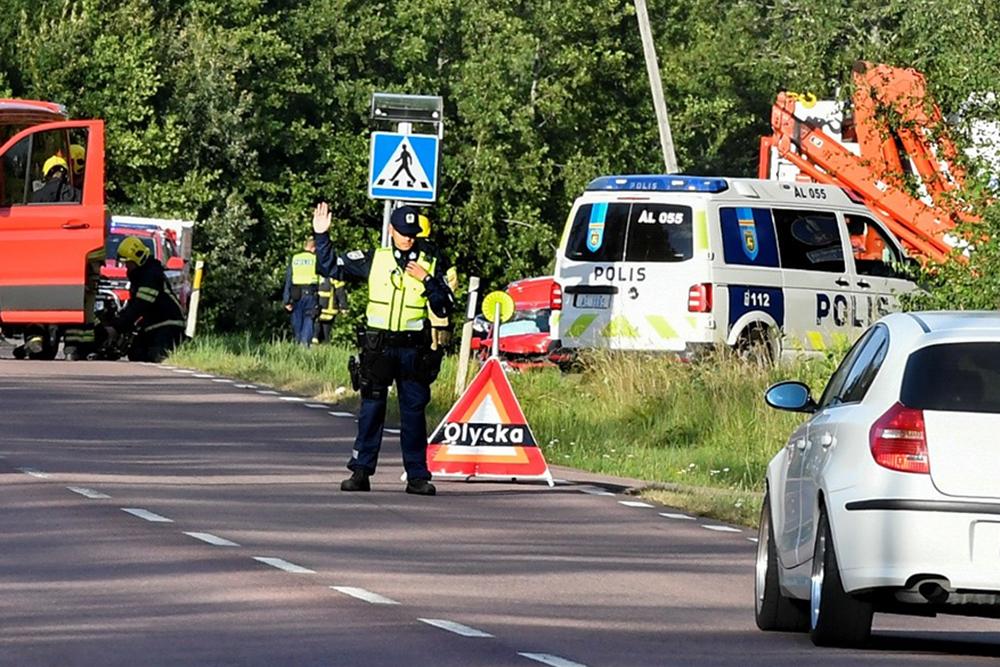 En polisman med tjänsteuniform står på vägen vid en olycksplats och dirigerar trafiken. Polismannen har sin högra hand i luften och handflatan riktat till den motkommande trafiken som ska stannas. I bakgrunden finns en polisbil och en brandbil och räddningspersonal som jobbar. 